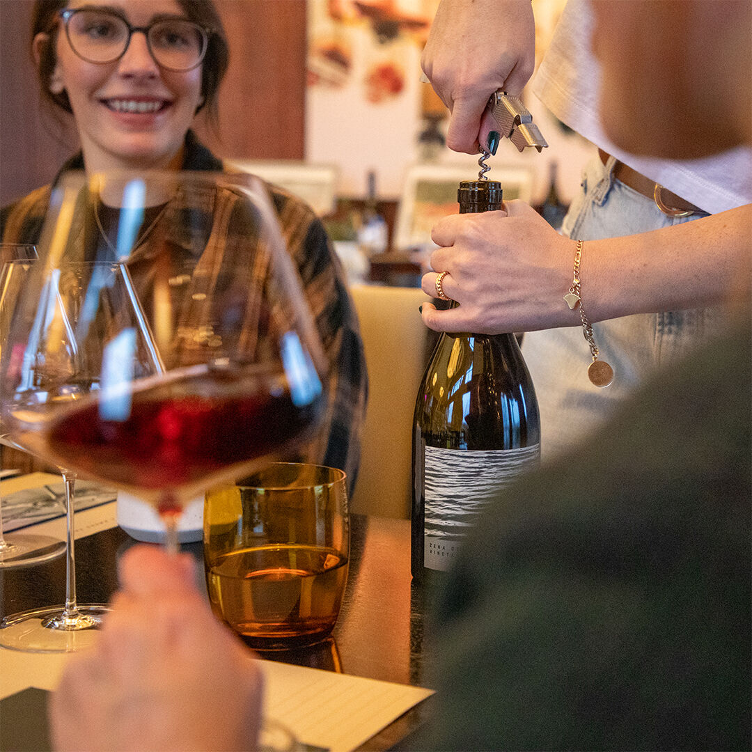 2 women chatting and swirling wine glasses as bottle is being opened by estate host 2 women chatting and swirling wine glasses as bottle is being opened by estate host